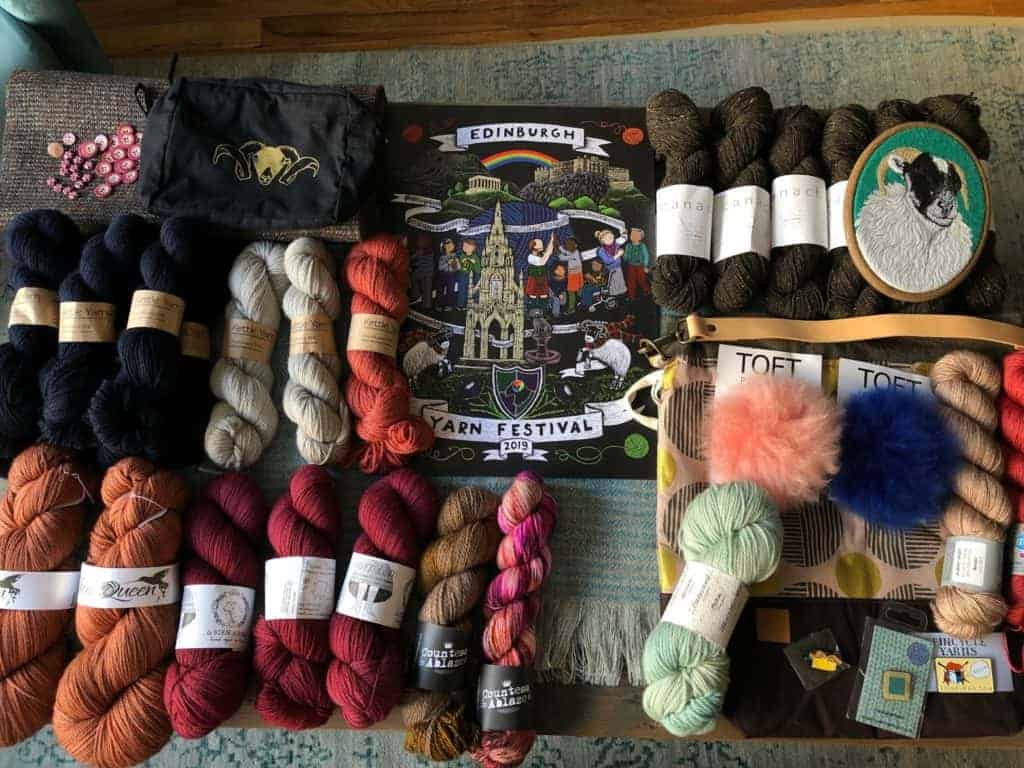 A collection of yarn, pompoms and buttons surrounds a poster for the Edinburgh Yarn Festival.