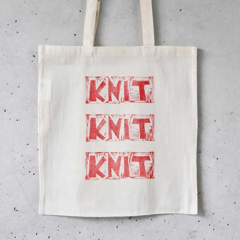 A cream tote bag with the words Knit Knit Knit in red.