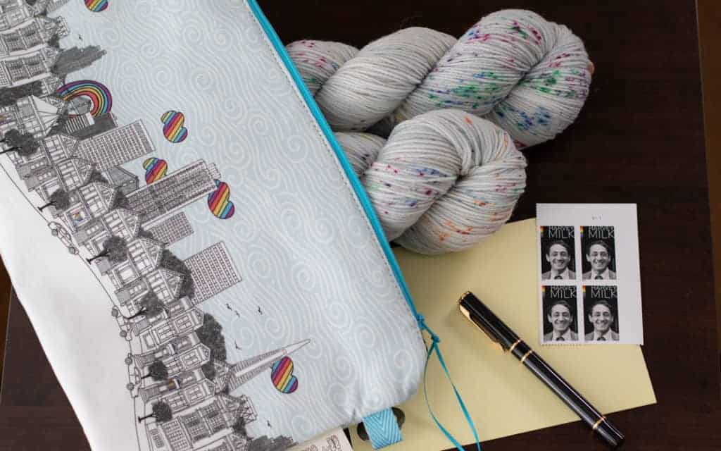 A project bag with a city skyline holds two skeins of gray and aqua speckled yarn