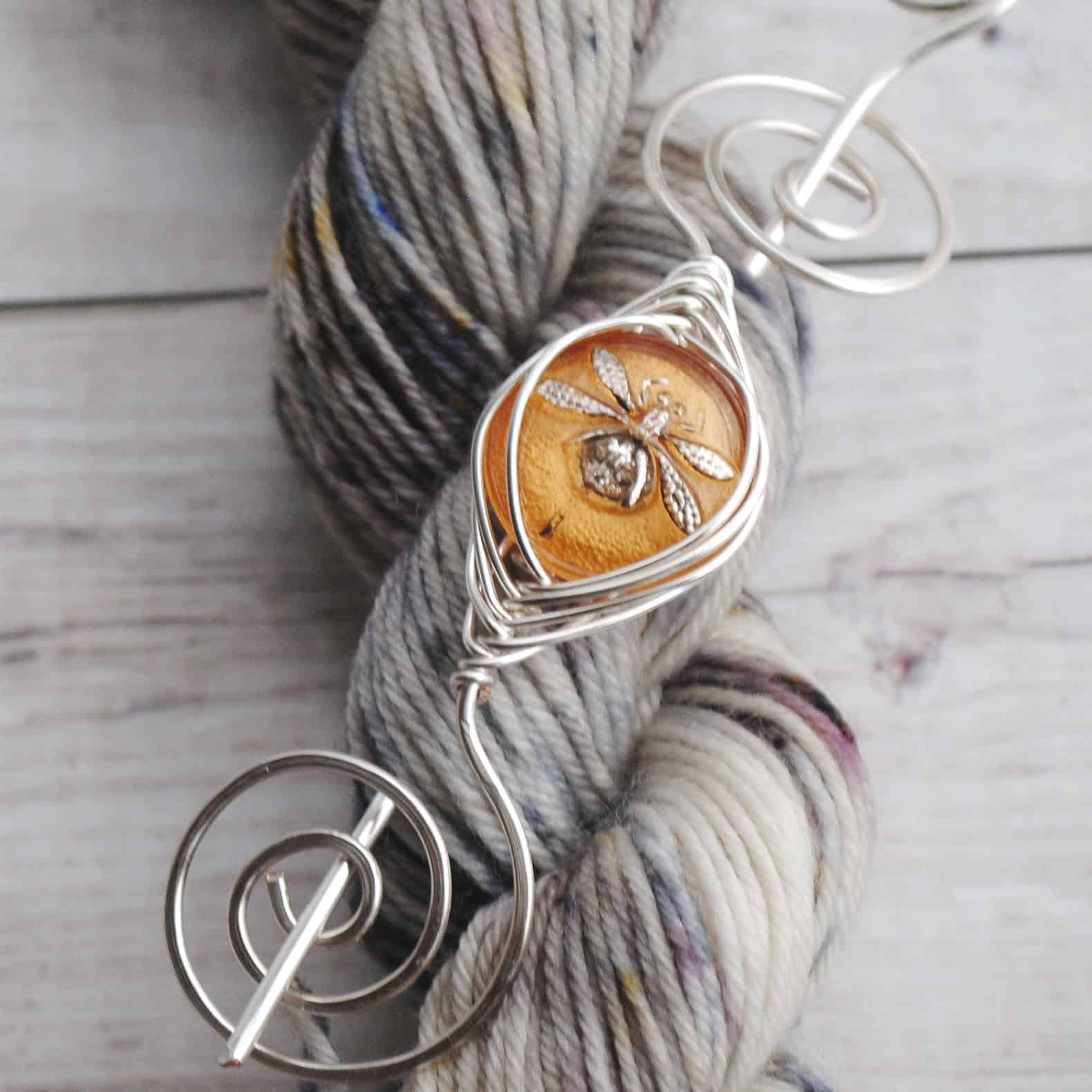 A dragonfly in amber in a silver shawl pin on a skein of gray yarn.
