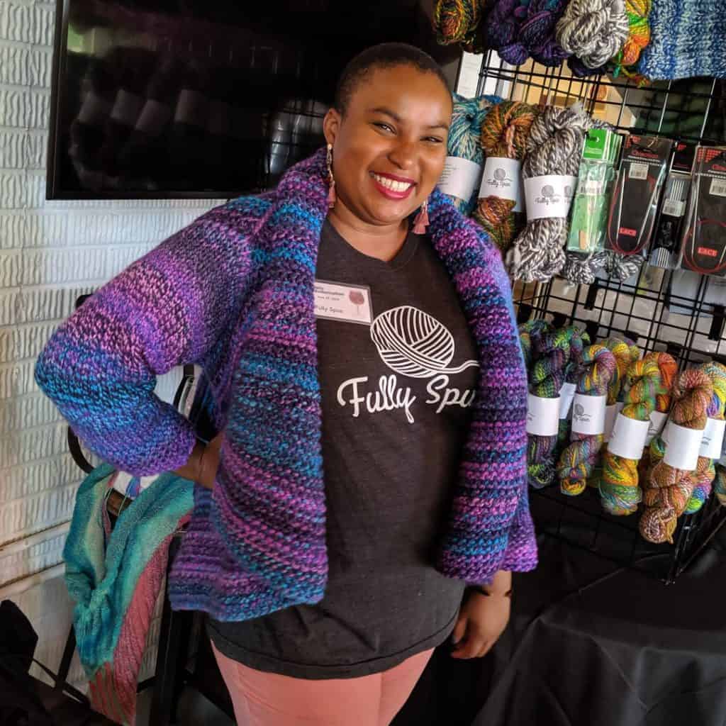 A black woman in a blue and purple cardigan smiles in front of a wall of colorful yarn.