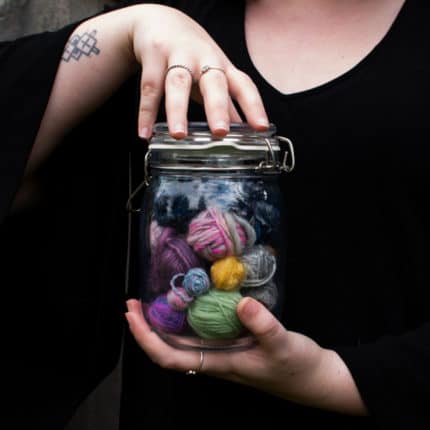 Hands holding a jar of brightly colored yarn balls.