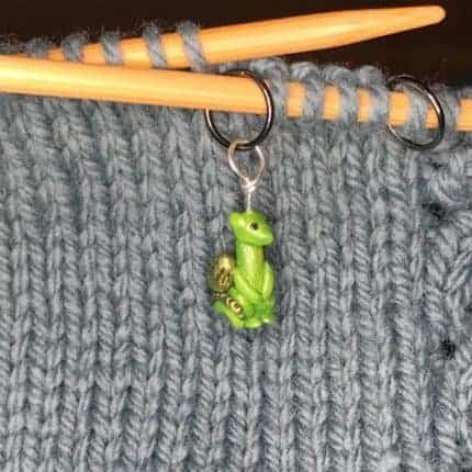 A green dragon stitch marker on wooden knitting needles.