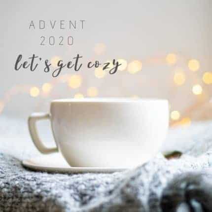 A white teacup sits in front of fairy lights on a handknit with the worlds Advent 2020 let's get cozy.