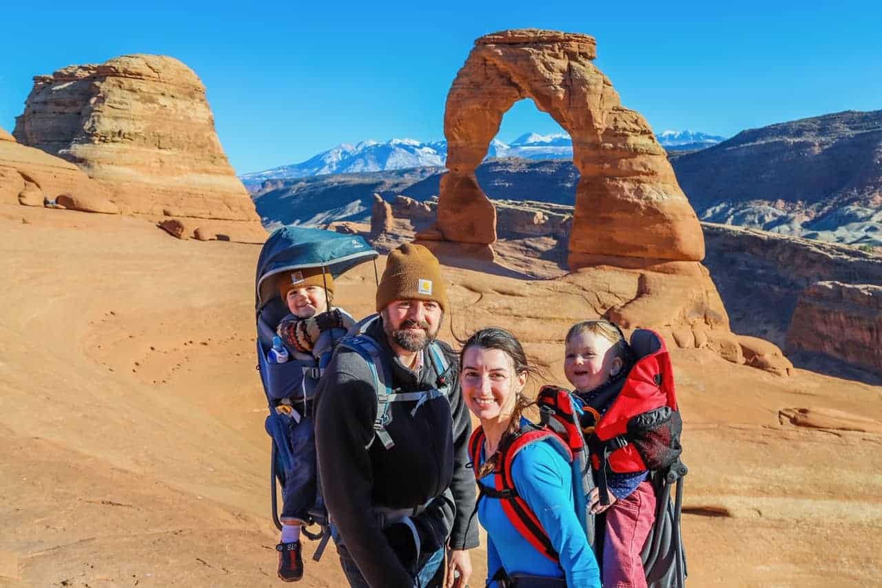 A mom and dad, each with a child on their back, pose in front of red rocks.