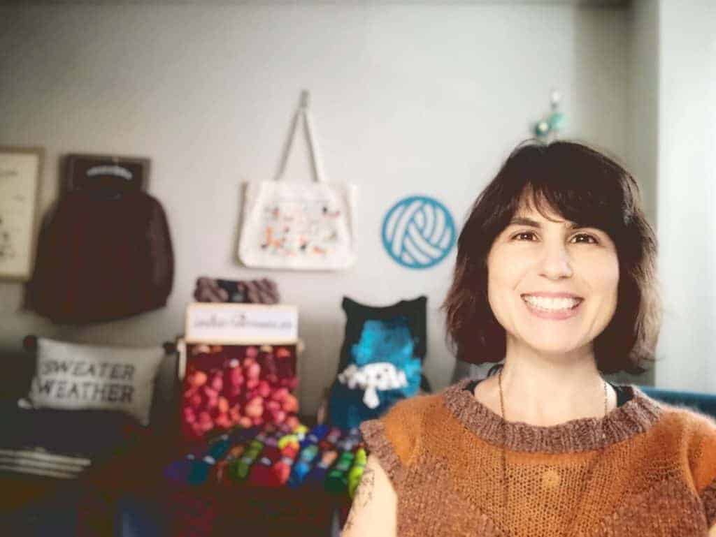 A white woman with short chin-length brown hair stands in front of a display of yarn beaming at the camera.