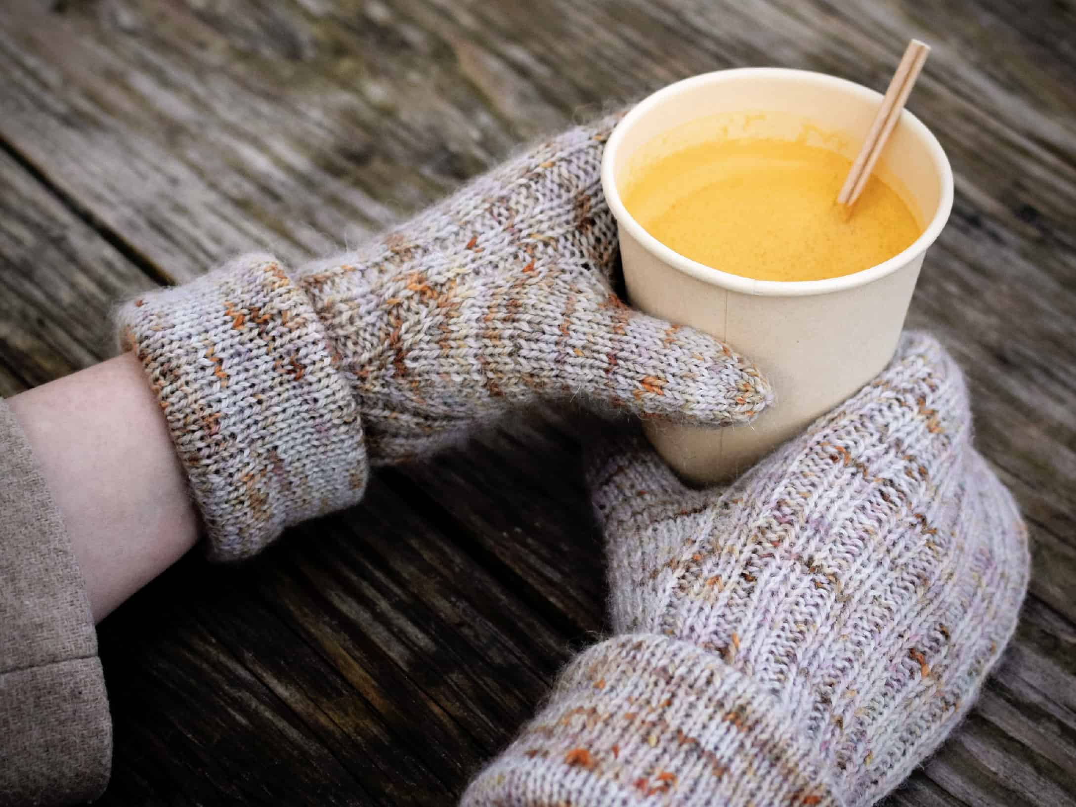 Hands in gray speckled mittens holding a golden beverage.