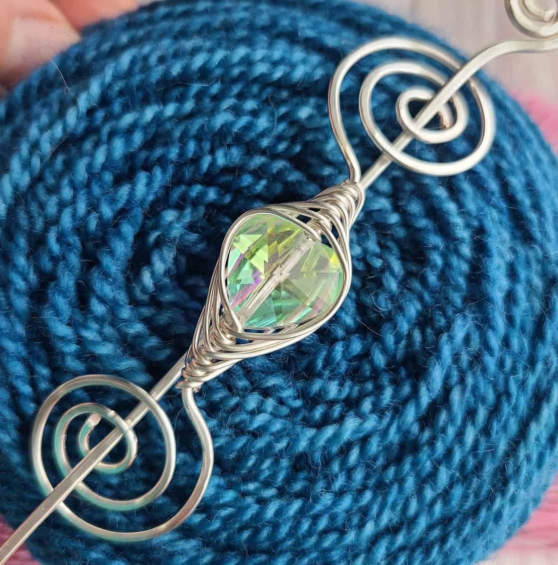 A silver shawl pin with a green stone on a cake of blue yarn.