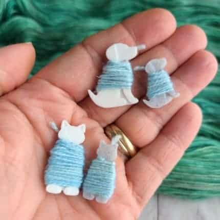 A hand holds white plastic dogs wrapped in light blue yarn.