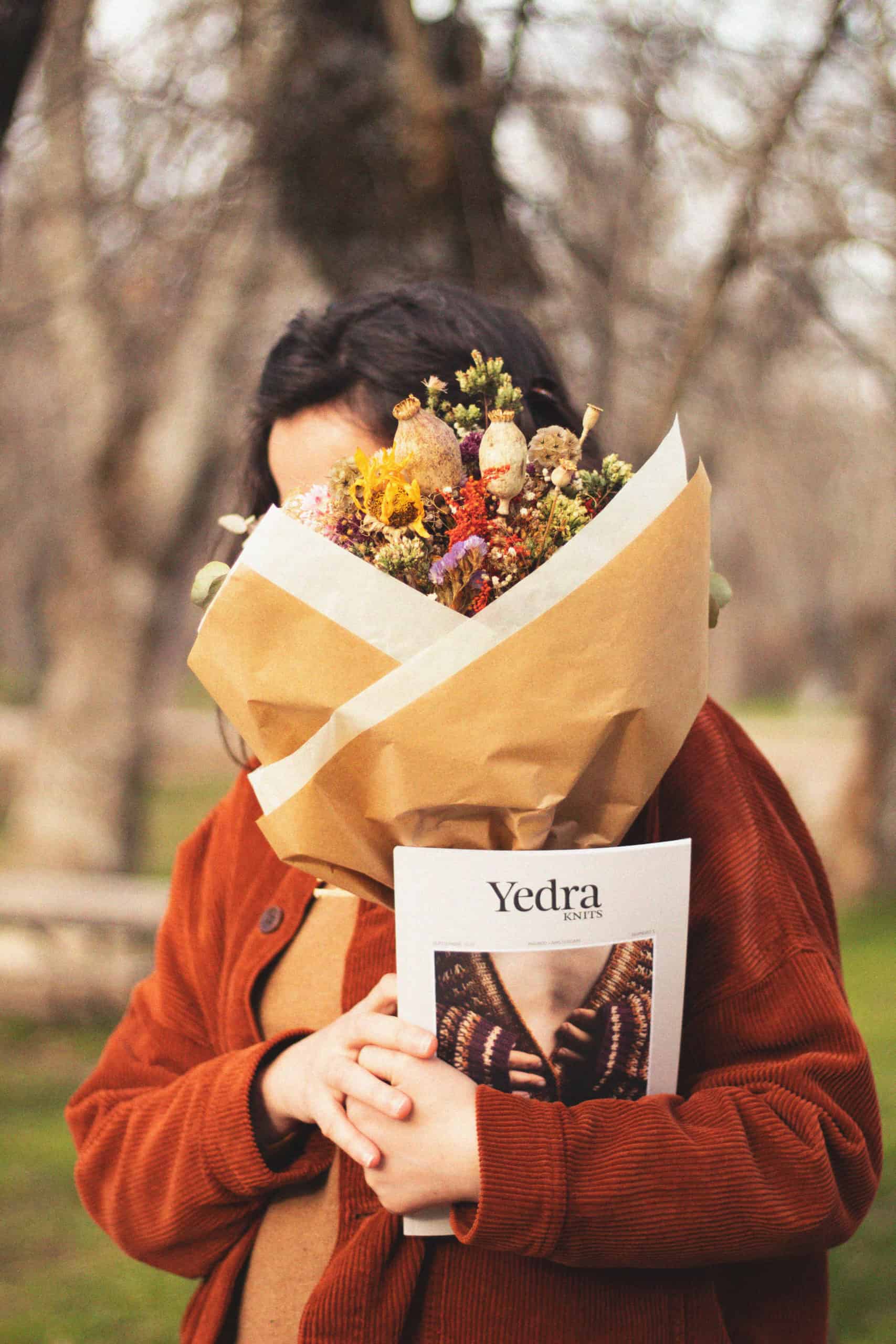 A person in an orange coat holding a bouquet of flowers in front of their face and a magazine called Yedra.