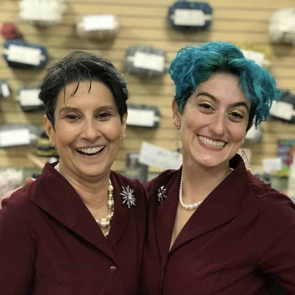 Two woman wearing purple blazers, one with teal hair, smile at the camera.