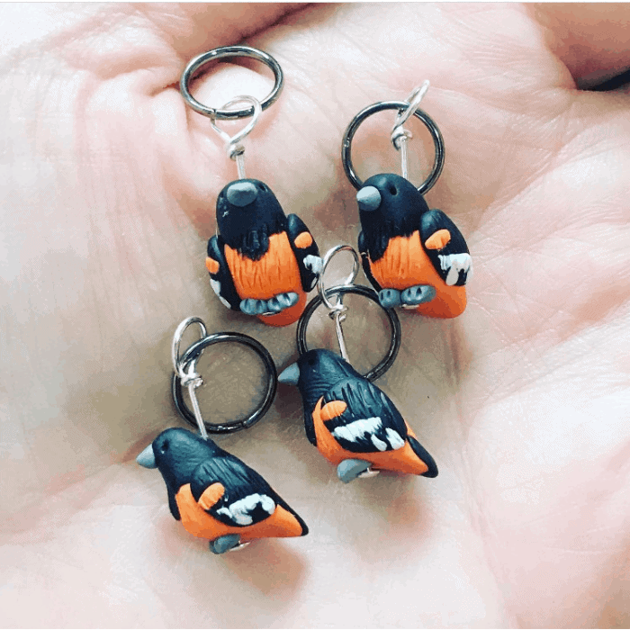Orange and blue bird stitch markers in a hand.