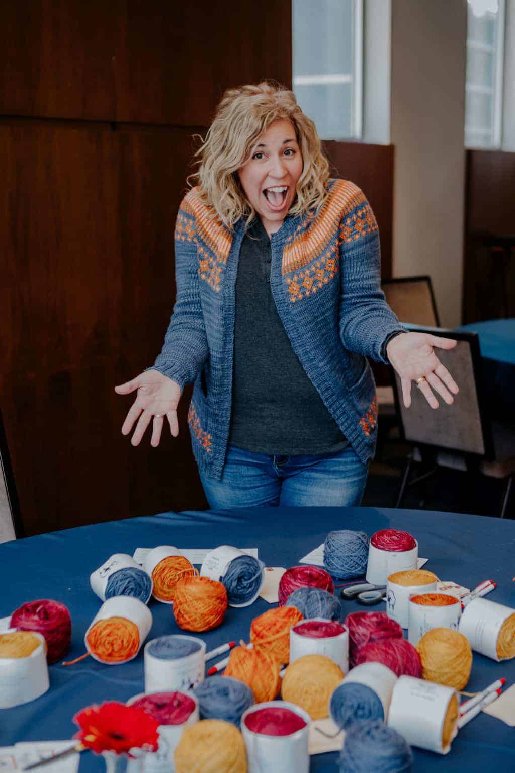 A woman wearing a blue and orange sweater stands over a table of colorful yarn.
