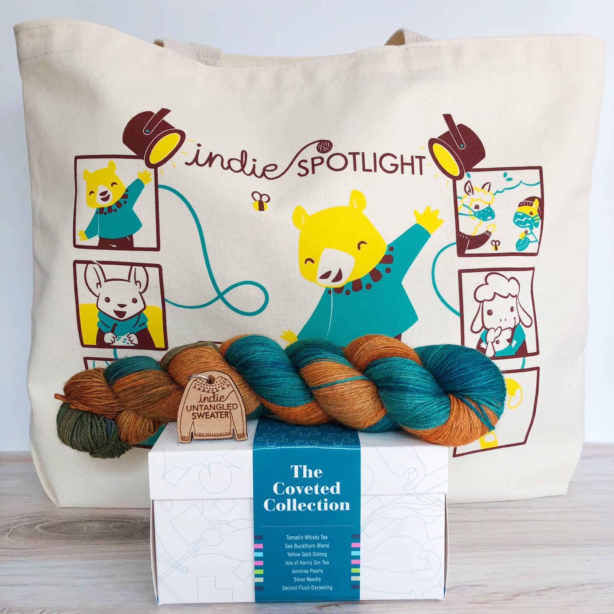 A tote bag with a gold bear, orange and teal yarn, a box of tea and a wooden sweater fob.