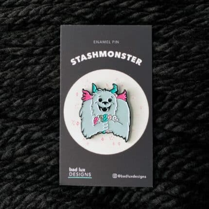 An enamel pin with a blue monster clutching skeins of pink and teal yarn.