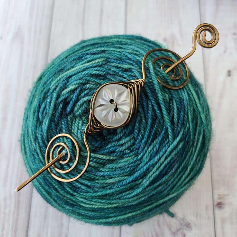 A spiral-edged pin with a mother of pearl button in the center sits atop a cake of green yarn.