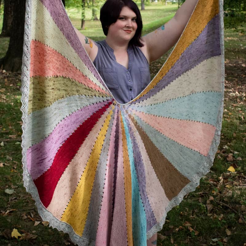 A light-skinned woman holds up a circular shawl with multicolored wedges.