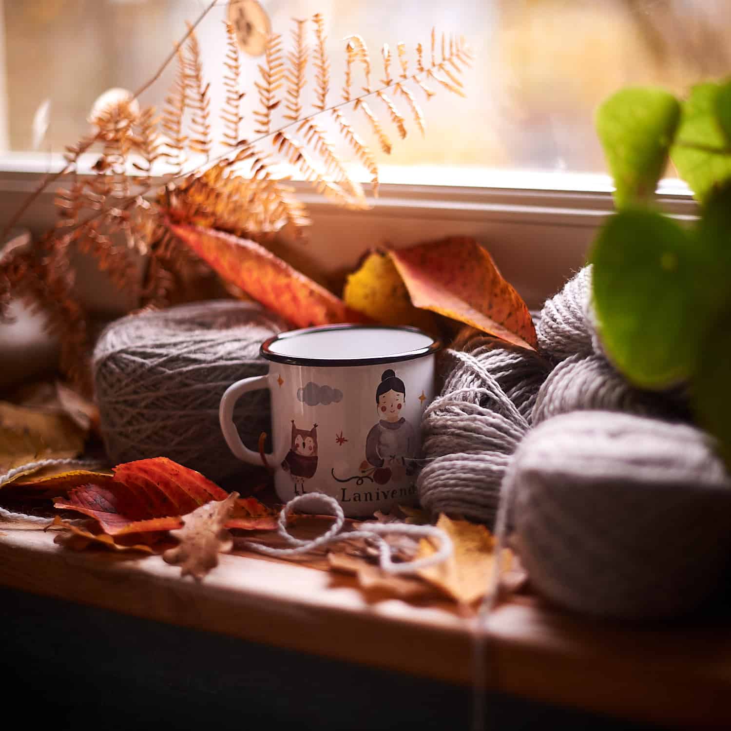 An illustrated mug sits by a window surrounded by gray yarn and red and orange fall leaves.