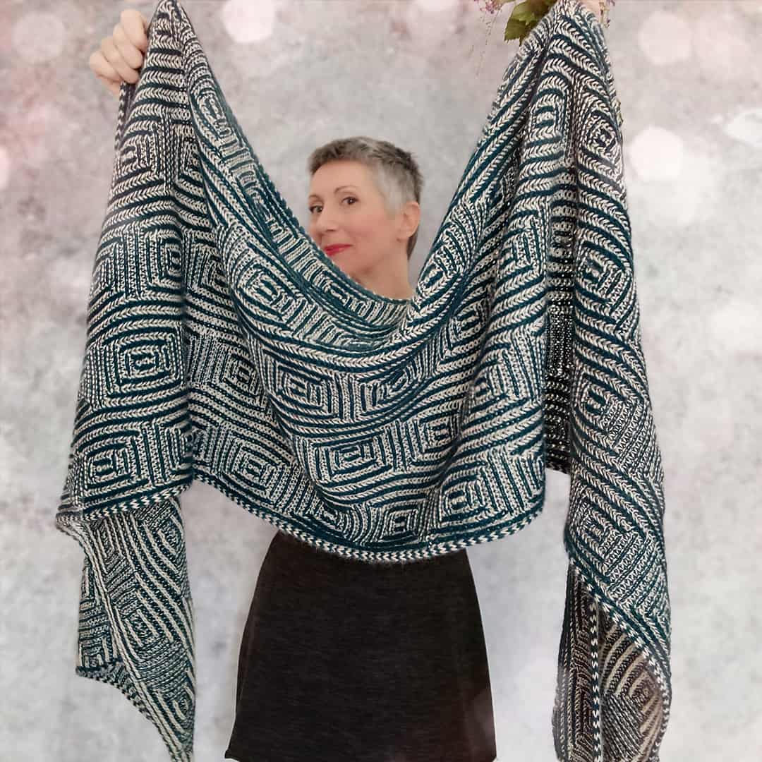 A white woman holds up a brioche shawl in shades of gray. 