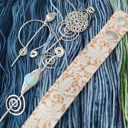 Silver swirled shawl pins and a cork band with a white floral pattern on loose skeins of blue hand-dyed yarn.