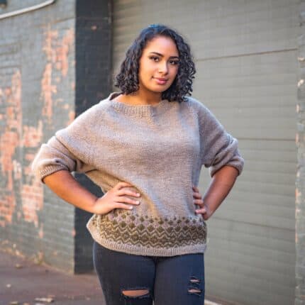 A brown-skinned woman wears a beige sweater with olive green colorwork at the hem.