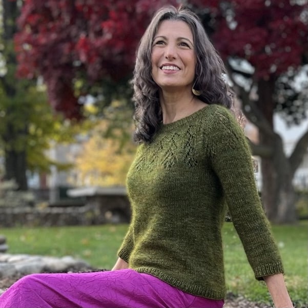 A woman wears a pink skirt and olive green handknit sweater.