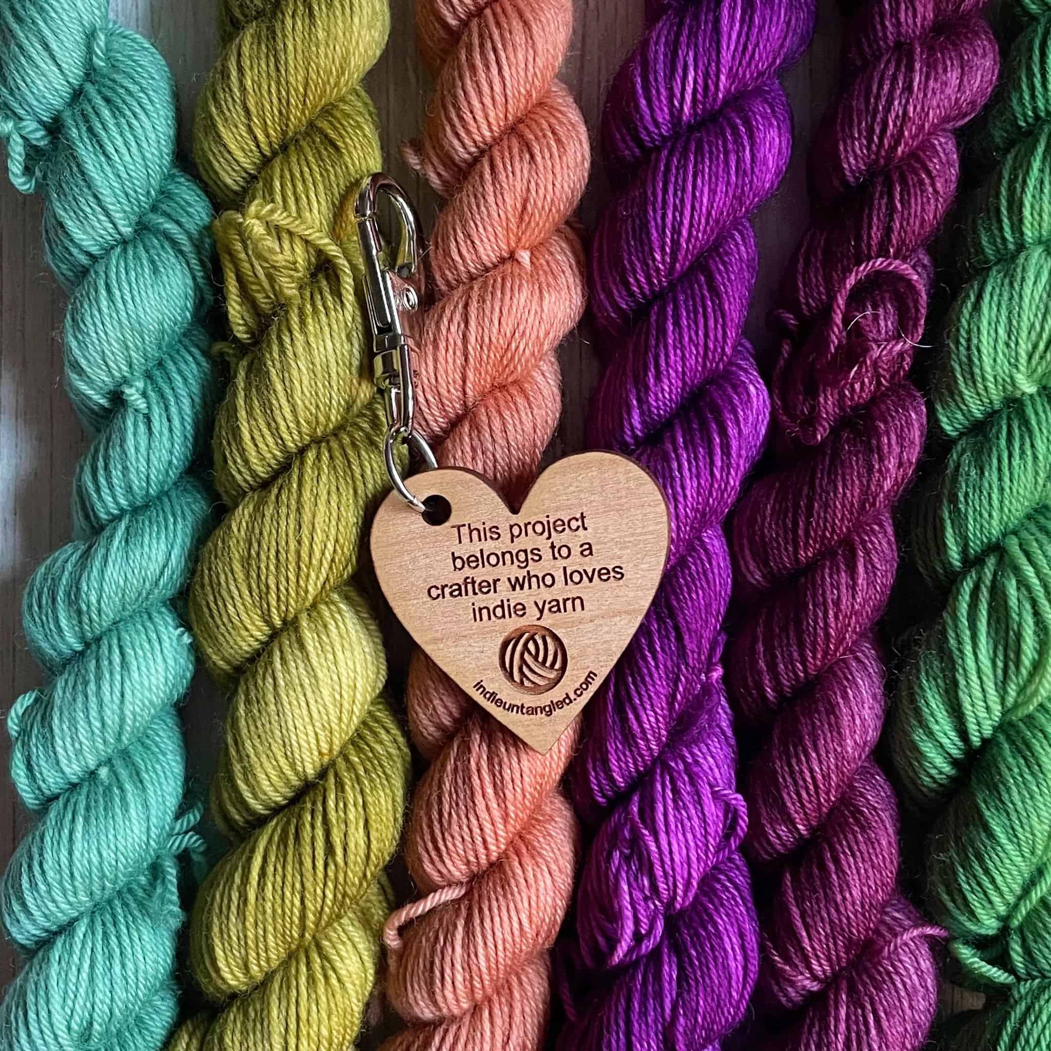 A wooden fob in the shape of a heart engraved with the words This project belongs to a crafter who loves indie yarn.