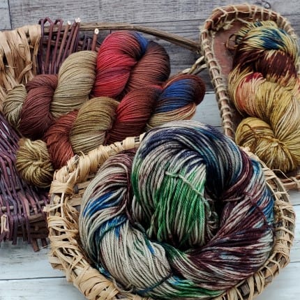 Four skeins of yarn in shades of brown, beige, and tan displayed in handwoven baskets. Splashes of red, blue, green, and darker browns across the various skeins.
