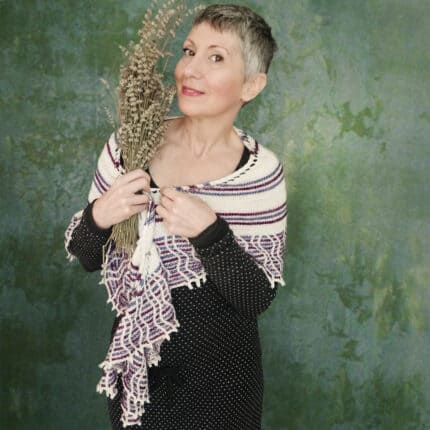 A light-skinned woman holding a dried lavender bouquet. She is wearing a striped shawl with a big multicolored border worked with slipped cables.