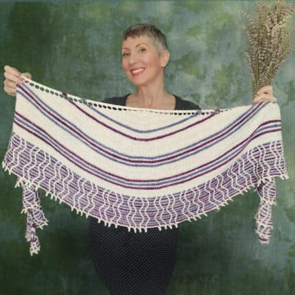 A light-skinned woman holding a crescent shawl open. The shawl is striped and has a big multicolored border worked with slipped cables.