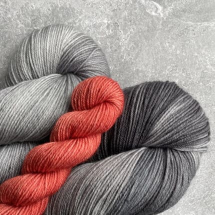 Two gray skeins of yarn, one light and one dark, with a small red mini skein.