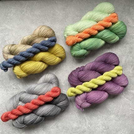 Four groupings of yarn lay on a gray surface, bathed in light. Each group contains two full skeins and one mini skein of different colors.