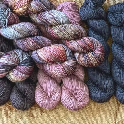 Three skeins of a purple, pink, blue variegated yarn with multicolored speckles on top of solid skeins in two different blues and dusty pink.