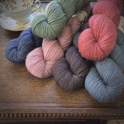 One skein each of dusty green, soft red, dusty pink, muted brown and two different blues on a wooden tray.