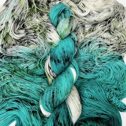 A white and teal fade skein with black speckles, laying on another skein.