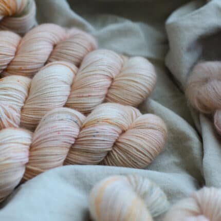 A pile of blush with light green and grey speckled yarn laying on a linen fabric.