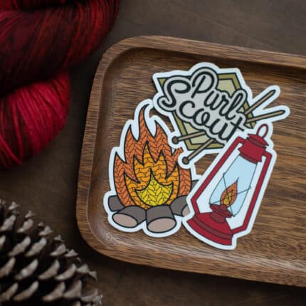 Three knitting stickers on a wooden tray including a campfire, lantern, and Purl Scouts logo.