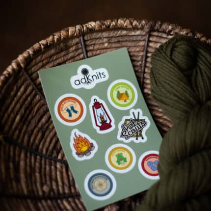 A sticker sheet of knitting-themed stickers in a basket with dark green yarn.