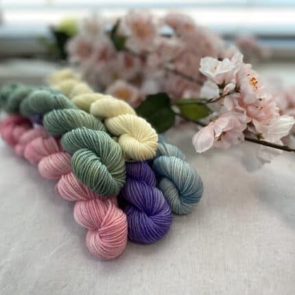 A stack of five pastel-colored mini skeins on a chair, with pale pink flowers in the back ground. Yarns are light green, cream, baby pink, lilac and sky blue.