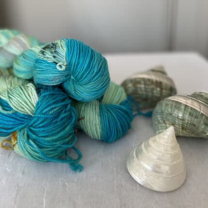 Three skeins of yarn in varying shades of light mint to turquoise blue with speckles. Three seashells are arranged around the skeins.