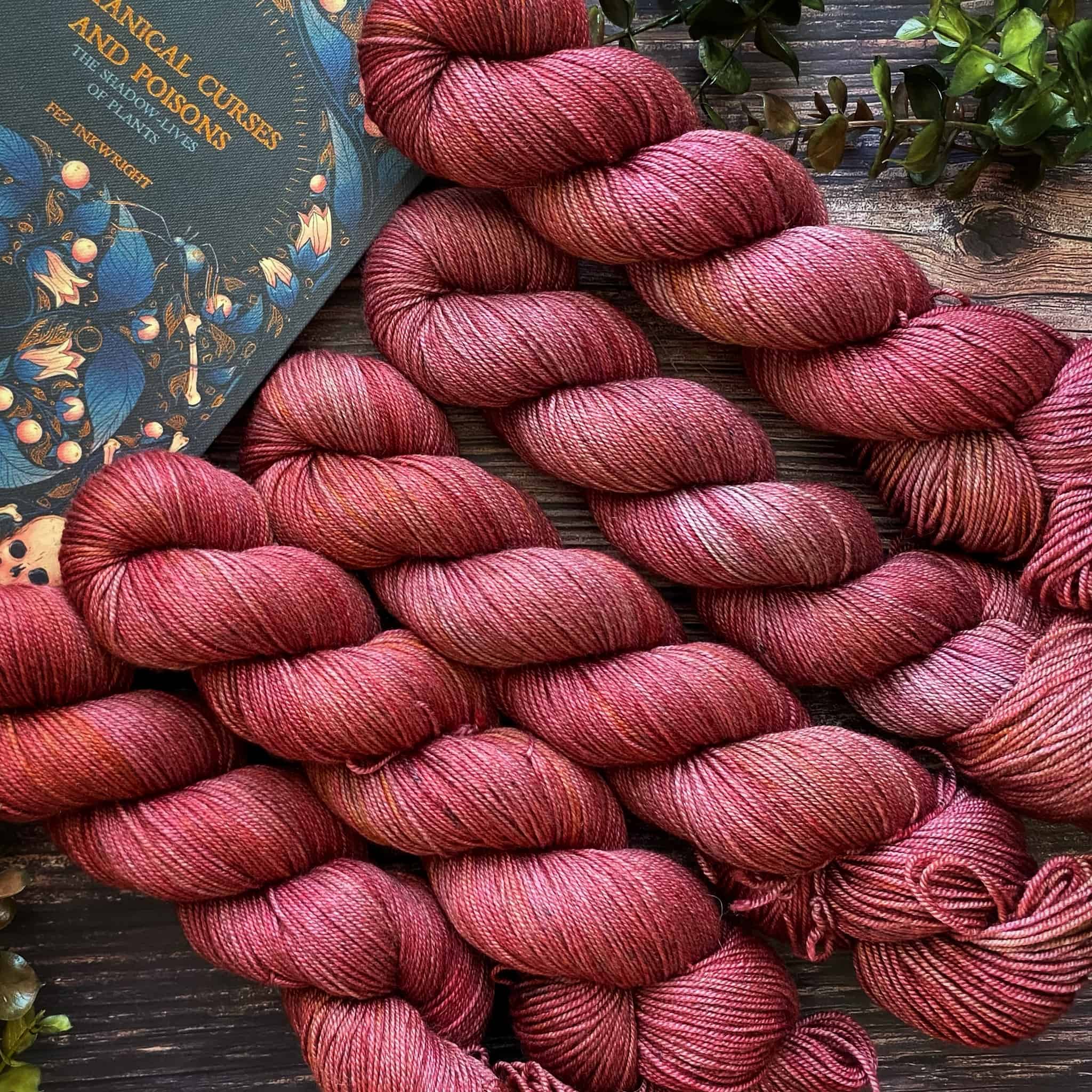 An angled view of five moody red skeins with hints of silver and brass surrounded by greenery and laying against a blue book with the title "Botanical Curses and Poisons."
