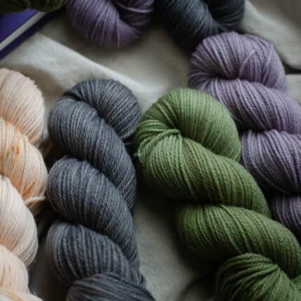 Four skeins of yarn, blush, grey, green and purple, laying on a cream linen fabric.