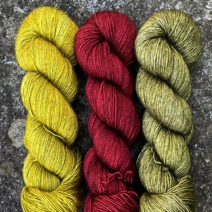 One skein each of mustard, dark red and olive green yarn on a stone background.