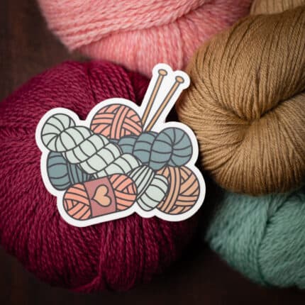 A sticker of yarn skeins and knitting needles sits on a pile of yarn.