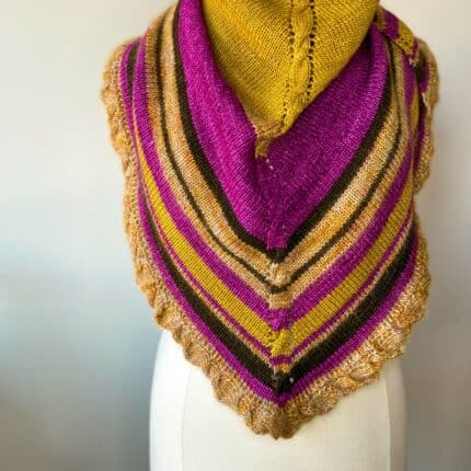 A knit shawl in gold, magenta and brown draped over a dress form showcasing a cable down the spine and along the border.