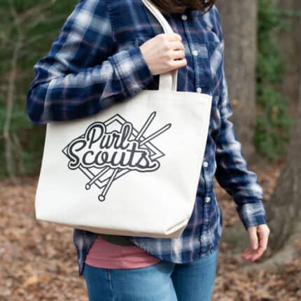A woman in a plaid shirt with a tote bag that says "Purl Scouts" over her shoulder.