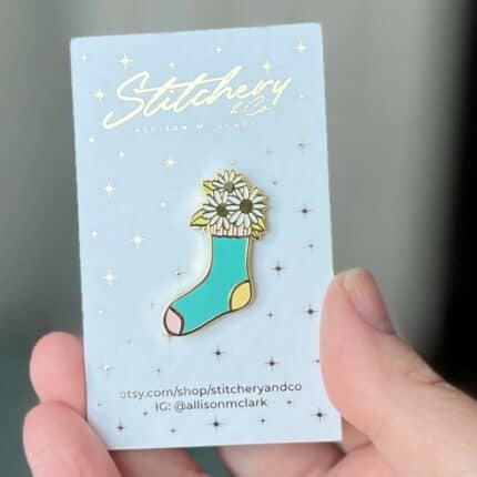 An enamel pin shaped like a sock with daisies inside it.