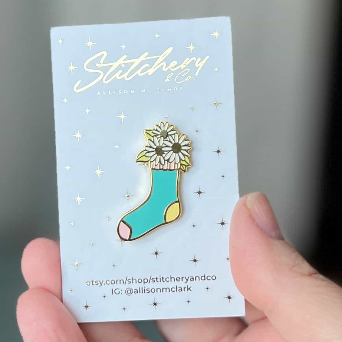 An enamel pin shaped like a sock with daisies inside it.