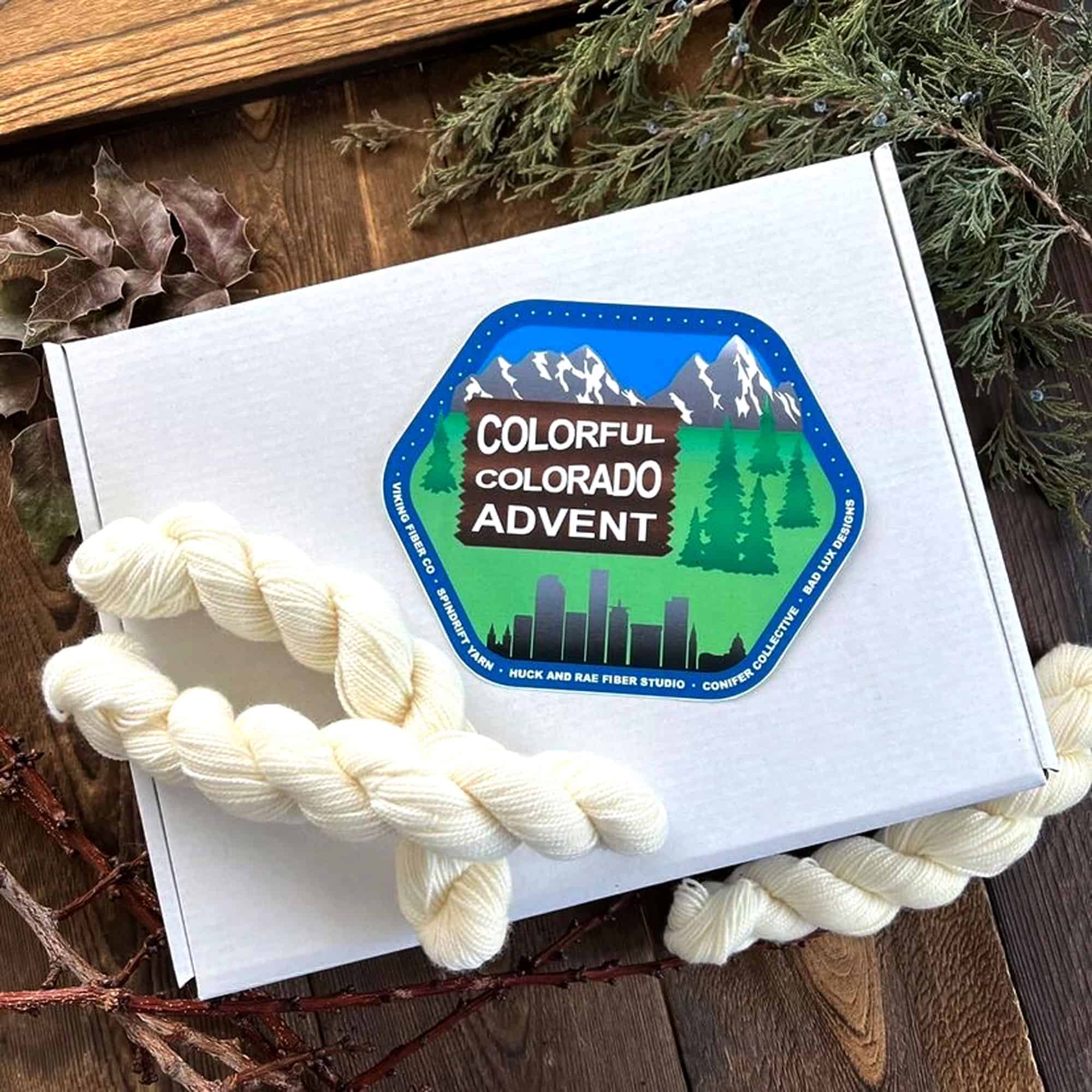 A white box surrounded by pine and holly branches with a logo on top of the box that reads Colorful Colorado Advent.