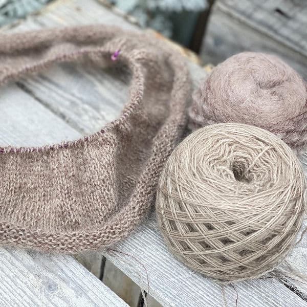 Two cakes of light brown yarn being knit into a tubular cowl.